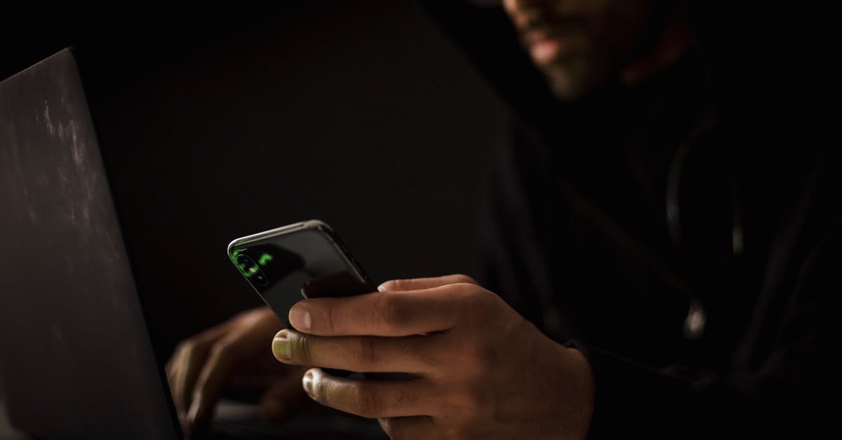 Crop anonymous ethnic male cyber spy with cellphone and netbook hacking system in evening